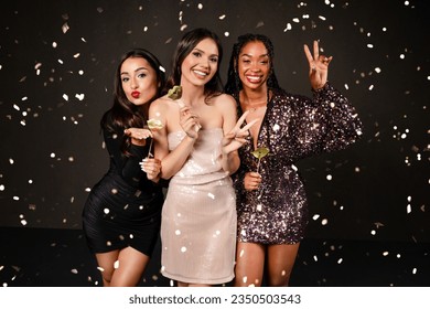 Cheerful attractive multicultural ladies in nice outfits posing together among falling confetti over black background, holding photo booth pops, grimacing and gesturing. New Year party