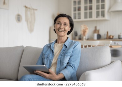 Cheerful attractive senior freelance woman using gadget at home, holding tablet computer, enjoying wireless domestic Internet connection, online app, web service, smiling at camera. Female portrait