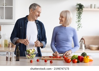 Cheerful Attractive Senior Man And Woman In Casual Outfits Cooking Together At Home, Making Delicious Vegetable Salad, Talking And Smiling, Kitchen Interior, Copy Space. Healthy Diet For Seniors