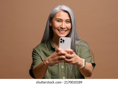 Cheerful Attractive Senior Woman Holding Smartphone, Enjoying Chatting In Social Networks, Shooting Someone, Smiling, Using Mobile App, Isolated On Brown