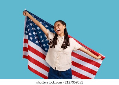 Cheerful attractive woman with black dreadlocks holding USA flag over shoulders and keeps eyes closed and smiling happily, wearing white shirt. Indoor studio shot isolated on blue background. - Powered by Shutterstock