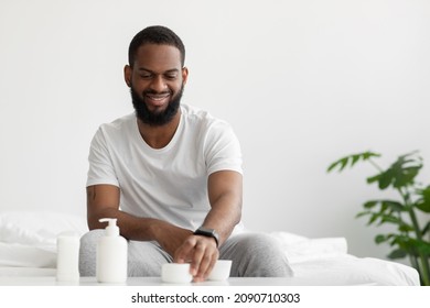 Cheerful Attractive Young African American Man With Beard Takes Jar Of Cream In Hand In Bedroom Interior On White Wall Background. Covid-19 Pandemic, Skin Care And Male Beauty At Home, Copy Space