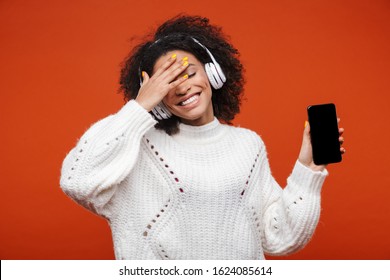 Cheerful attractive young african woman wearing wireless headphones standing isolated over red background, showing blank screen mobile phone