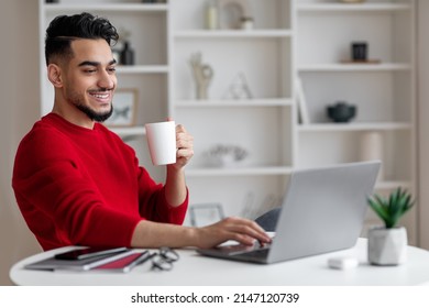 Cheerful Attractive Young Arab Businessman With Beard In Red Clothes Drinks Coffee And Types On Laptop At Workplace In Home Office Interior. Break From Work Remotely, Rest And Relax During Covid-19