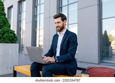 Cheerful Attractive Young Caucasian Guy Manager With Beard In Suit Typing On Laptop, Sits On Bench Near Modern Office Building. Successful Business, Social Distance, Businessman Have Meeting Outdoor