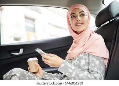 Cheerful Attractive Young Muslim Businesswoman In Beautiful Headscarf Sitting On Backseat And Using Mobile App For Taxi Ride