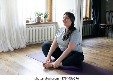 Cheerful attractive young overweight woman in activewear choosing healthy lifestyle, sitting on mat with hands on bare feet, doing butterfly yoga exercise, stretching thighs. Body shape and activity