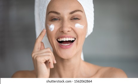 Cheerful Attractive Young Woman Apply Facial Creme Laughing Looking At Camera, Happy Lady Wrap Towel On Head Putting Cream Lifting Hydrating Moisturizing On Face Skin Care Concept, Close Up Portrait