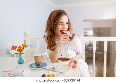 Cheerful attractive young woman with long curly hair sitting and eating heart shaped cookies 