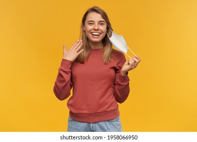Cheerful attractive young woman taking off her virus protective mask on face against coronavirus and smiling over yellow background