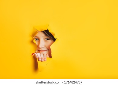 Cheerful Baby Girl Face Looks Out Through Torn Hole In Yellow Paper Wall. Peek Through Hole. Discover Something New. Looking Beyond. Aside Place For Text Information, Advertising, Positive News.