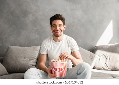 Cheerful Bachelor Sitting On Sofa In Gray Apartment And Laughing While Watching Tv With Pop Corn In Hands