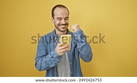 A cheerful bald hispanic man with a beard celebrating a message on his phone against a yellow background.