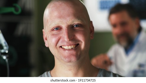 Cheerful bald man with cancer smiling and looking at camera after hearing remission news in oncologist office in hospital