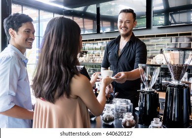 Cheerful bartender serving coffee over the bar counter to a young female customer standing next to her boyfriend in the cafeteria - Powered by Shutterstock