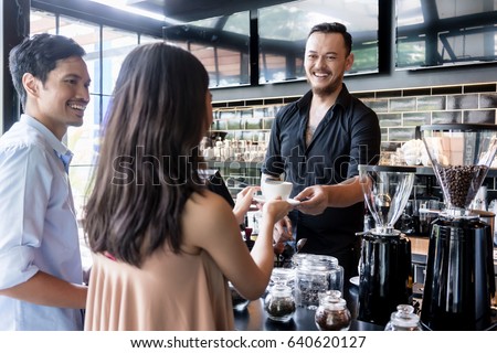 Cheerful bartender serving coffee over the bar counter to a young female customer standing next to her boyfriend in the cafeteria