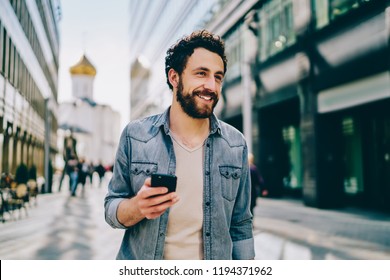 Cheerful Bearded 30s Young Man Dressed In Denim Wear Looking Away While Holding Smartphone Using 4G Internet Connection. Happy Hipster Guy Holding Mobile Phone While Walking Outdoors On City Street