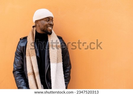 Cheerful bearded African American male wearing leather jacket scarf and hat standing against light orange wall and looking away with happy smile