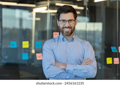 A cheerful, bearded businessman stands confidently in a well-lit office, arms crossed, with sticky notes on the glass background. - Powered by Shutterstock