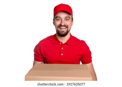 Cheerful bearded delivery man in red uniform and cap looking at camera with friendly smile while carrying big cardboard box against white background