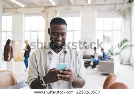 Cheerful bearded ethnic male worker in shirt sitting on table and texting message via mobile phone during workday with diverse colleagues in modern office