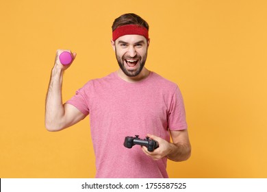 Cheerful Bearded Fitness Sporty Guy 20s Sportsman In Headband T-shirt In Home Gym Isolated On Yellow Background. Workout Sport Motivation Concept. Doing Exercise With Dumbbell Play Game With Joystick