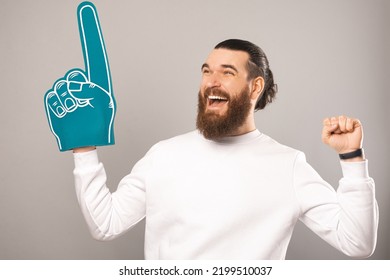 Cheerful bearded guy is celebrating victory while wearing foam fan glove. Studio shot over light grey background. - Powered by Shutterstock