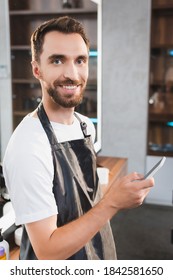 cheerful,  bearded hairstylist looking at camera while chatting on smartphone
