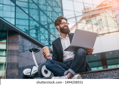 Cheerful Bearded Male Entrepreneur With Modern Laptop And Takeaway Coffee Smiling Away While Sitting On Stairs With Electric Transport Outside Building On City Street