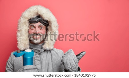 Cheerful bearded male skier with happy expression enjoys winter recreation dressed in warm jacket gloves covered with snow drinks hot beverage from thermos goes in for extreme sport in mountains