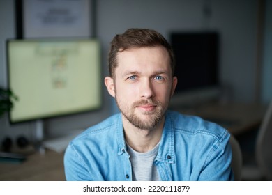 Cheerful Bearded Male Software Engineer Sitting At Working Place In Office Looking At Camera. Young Handsome Man In Denim Shirt, Positive Emotion With Computer Monitor On Background