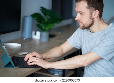 Cheerful Bearded Male Software Engineer Sitting At Working Place In Office Hardly Working Using Laptop. Young Handsome Man In Grey T-shirt, Positive Emotion With Computer Monitor On Background