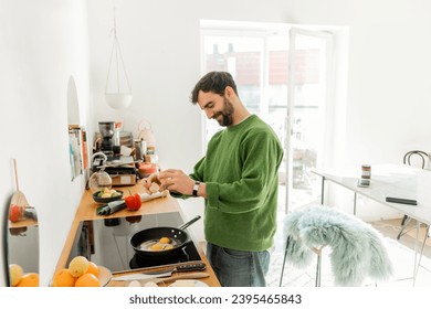 Cheerful bearded man in casual clothes pouring egg on frying pan while cooking breakfast - Shutterstock ID 2395465843