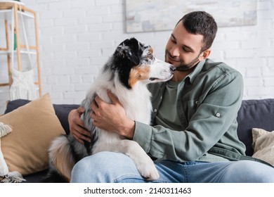 Cheerful Bearded Man Cuddling Australian Shepherd Dog On Couch