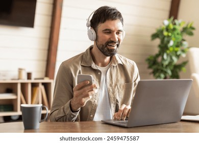 A cheerful bearded man is engaged in multitasking in a well-lit room; he uses smartphone and laptop simultaneously while wearing headphones. The setting suggests casual work-from-home environment - Powered by Shutterstock