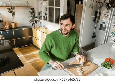 Cheerful bearded man in green jumper cutting fresh leek and looking away while cooking at home - Shutterstock ID 2395465763