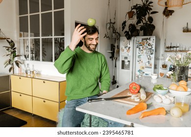 Cheerful and bearded man in green jumper balancing with apple on head in modern kitchen - Shutterstock ID 2395465781
