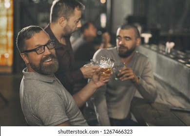 Cheerful Bearded Man Holding Crystal Glass Of Whisky, Looking At Camera And Smiling. Clients Of Bar Sitting At Counter Behind, Communicating And Drinking Elite Alcohol.