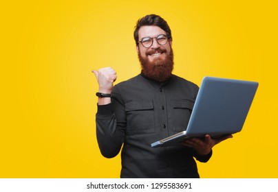 Cheerful Bearded Man Is Holding Laptop And Poiting Bkack, Looking At Camera Over Yellow Background