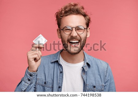 Cheerful bearded man holds condom, being glad to have unforgettable night with woman, wants to repeat once again. Positive stylish man promotes healthy lifestyle and relationship, shows how protect