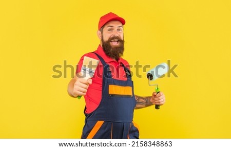 cheerful bearded man housepainter in work clothes hold paint roller and brush