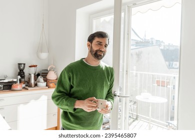 Cheerful bearded man in jumper holding cup of coffee and looking at camera - Shutterstock ID 2395465807