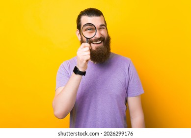 Cheerful bearded man is looking through a magnifying glass over yellow background in a studio.