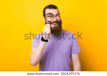Cheerful bearded man is looking through a magnifying glass over yellow background in a studio.