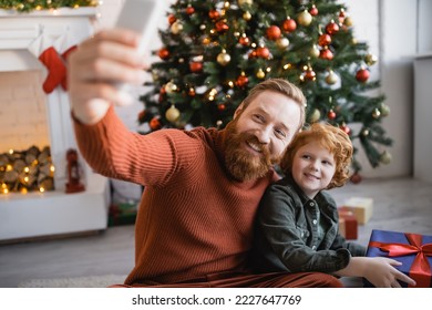 cheerful bearded man taking selfie with redhead son holding gift box near Christmas tree - Powered by Shutterstock