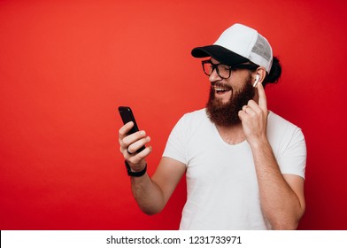 Cheerful Bearded Man Using Airpods And Looking At Phone Over Red Wall