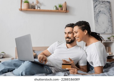 cheerful bearded man using laptop near african american woman with credit card on bed at home - Powered by Shutterstock