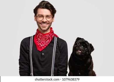 Cheerful bearded young male with trendy hairdo, wears stylish shirt and red bandana and his favourite black pug dog who looks with curious expression, being satisfied after good walk outdoor