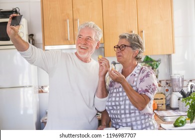 Cheerful beautiful aged senior couple working together in daily activity in the kitchen making food for lunch. The man take selfie to both to have fun and smile. matures using technology and internet - Powered by Shutterstock