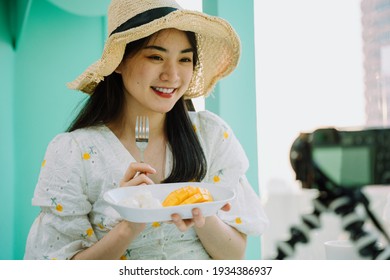 Cheerful beautiful asian thai woman wearing hat holding plaate of mango sticky rice and fork in front of camera. Doing food vlog.